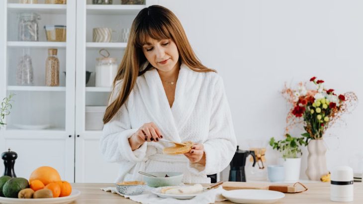 A woman in a bathrobe preparing breakfast in a home kitchen.